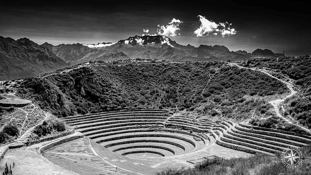 Black and white photo of Incan terraces at Moray with a snow-capped mountain and drifting clouds in the background.