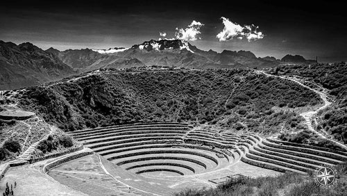 Black and white photo of Incan terraces at Moray with a snow-capped mountain and drifting clouds in the background.