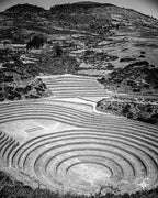 Black and white photograph of Incan terraces expanding outward and up a mountain, captured from a dramatic angle.