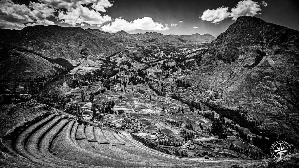 A bird’s-eye view of ancient Incan terraces carved into a steep mountain slope, descending toward a valley where modern vil