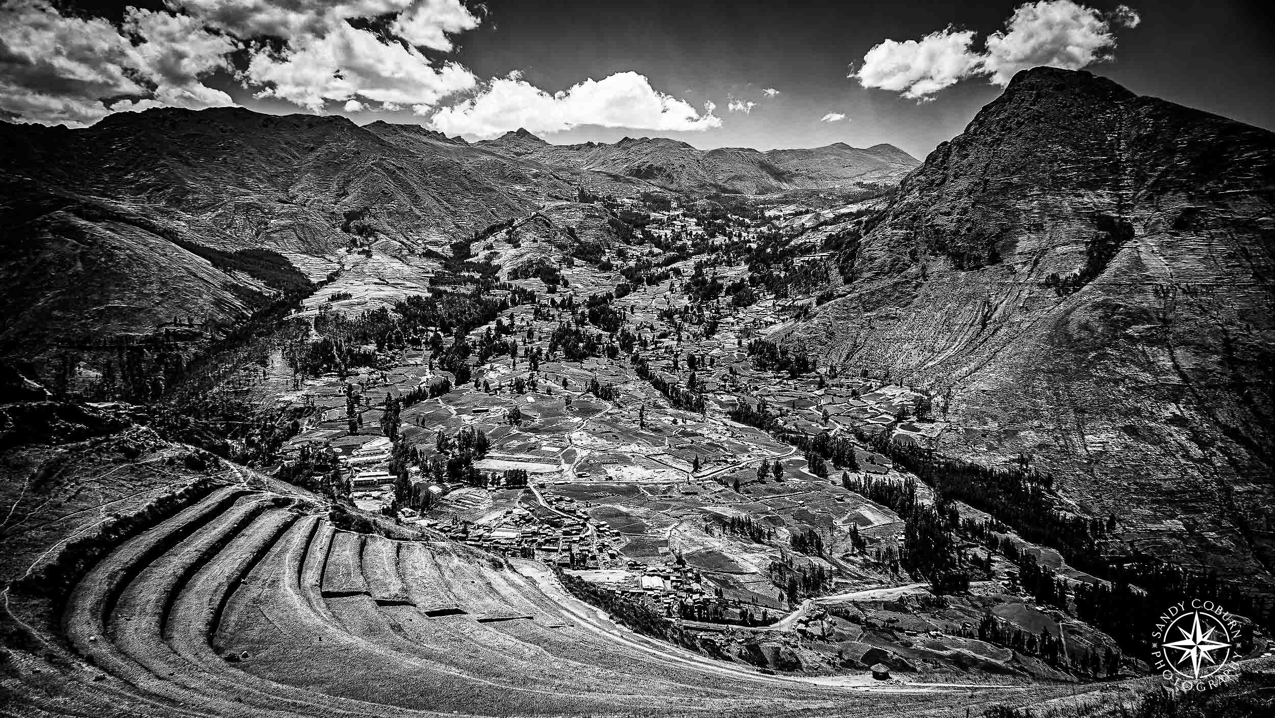 A bird’s-eye view of ancient Incan terraces carved into a steep mountain slope, descending toward a valley where modern vil