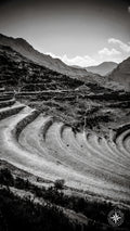 A dramatic black and white photograph of Incan terraces sweeping in a curve across the frame, with shadowed ridges, distant m