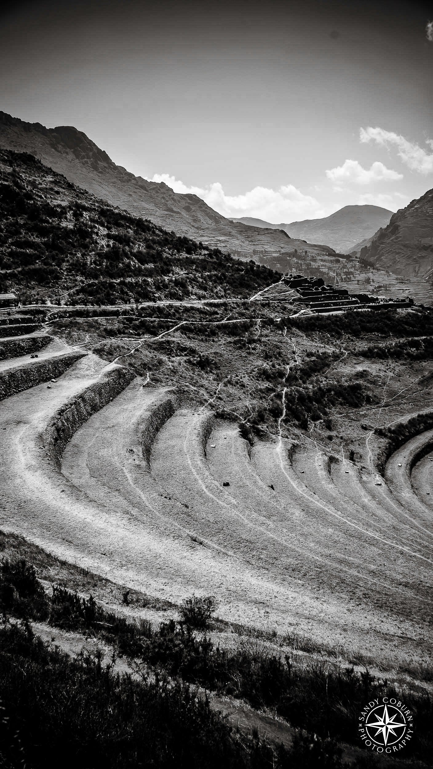 A dramatic black and white photograph of Incan terraces sweeping in a curve across the frame, with shadowed ridges, distant m