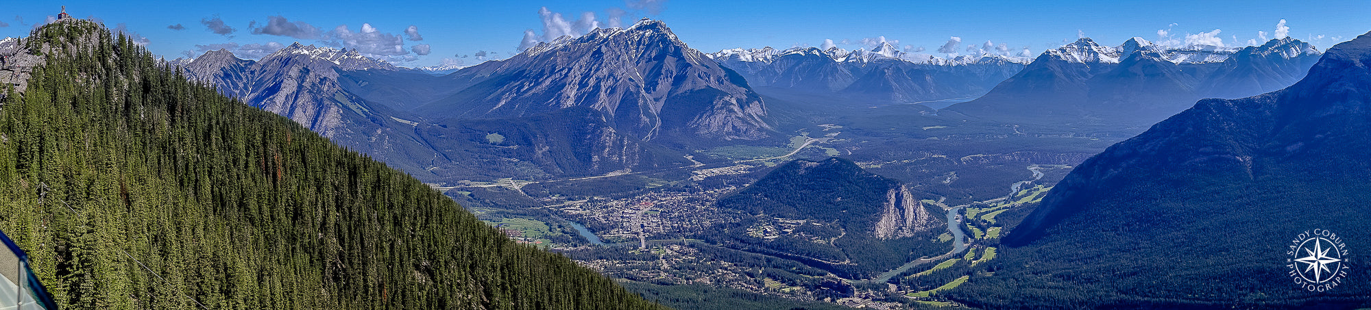 Aerial view from Sulphur Mountain showing a lush green valley surrounded by shadowy black and gray mountain ridges, under a c