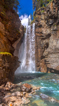 A powerful waterfall framed by rust-colored canyon walls plunges into a glowing turquoise pool along the Ink Blots Trail in B