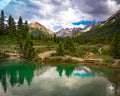 Teal and green glacial pool reflecting pine trees and mountains beneath a storm-filled sky in Banff National Park.
