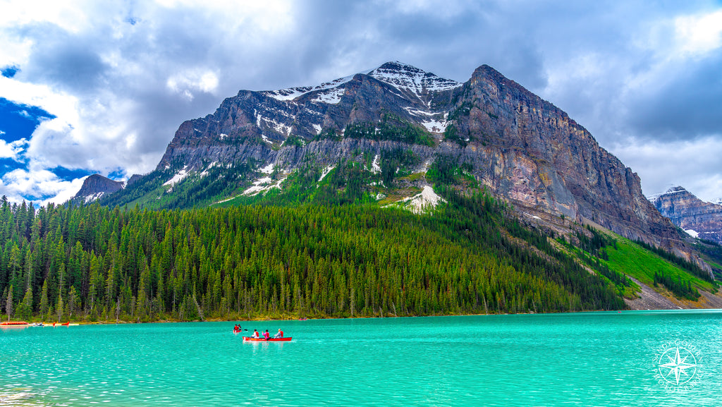 A red canoe paddles across the vibrant teal waters of Lake Louise, framed by evergreen trees, granite cliffs, and brooding pr