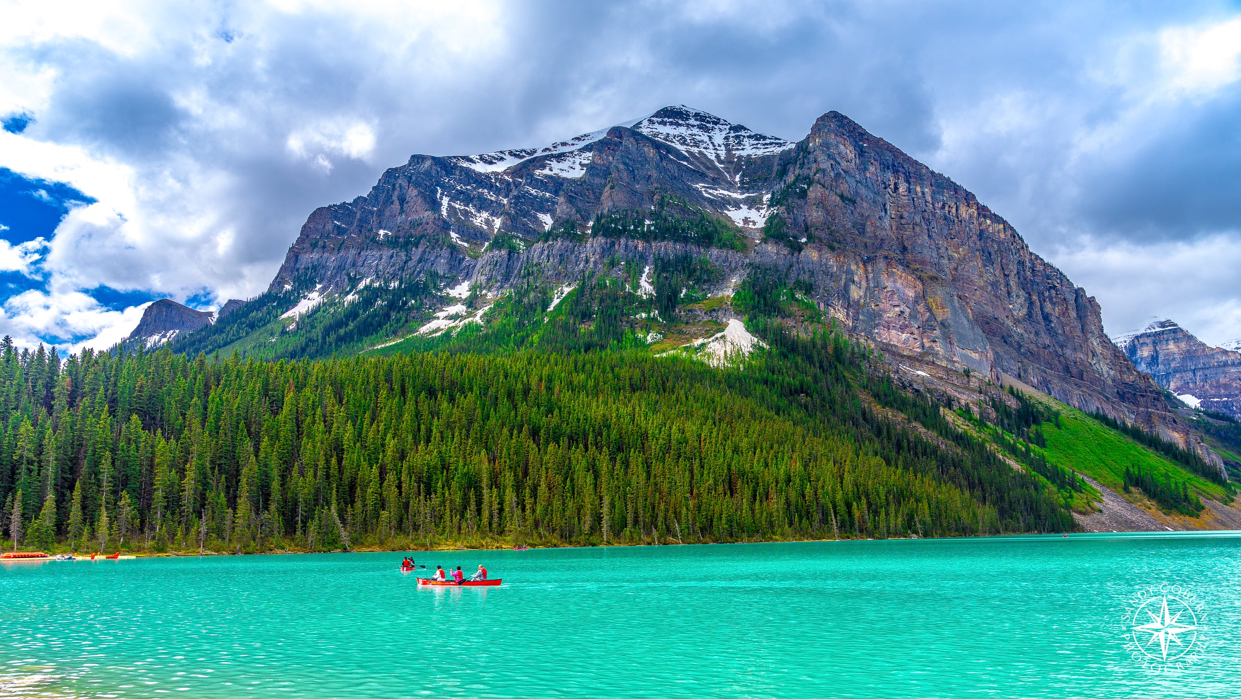 A red canoe paddles across the vibrant teal waters of Lake Louise, framed by evergreen trees, granite cliffs, and brooding pr