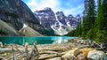 Snow-capped granite peaks tower over the dark blue-green waters of Lake Moraine, with driftwood gathered in the lower left co