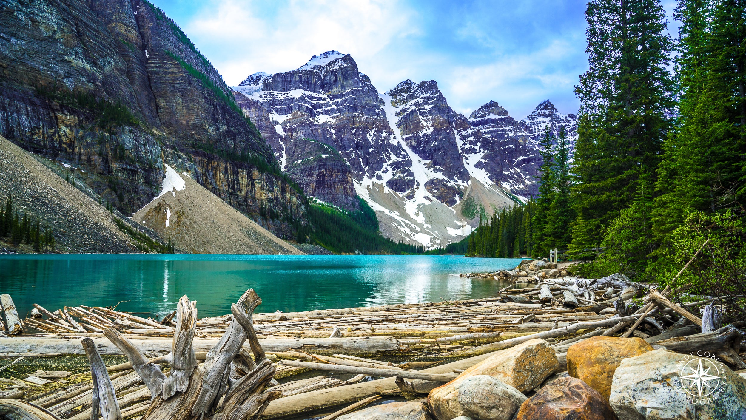 Snow-capped granite peaks tower over the dark blue-green waters of Lake Moraine, with driftwood gathered in the lower left co