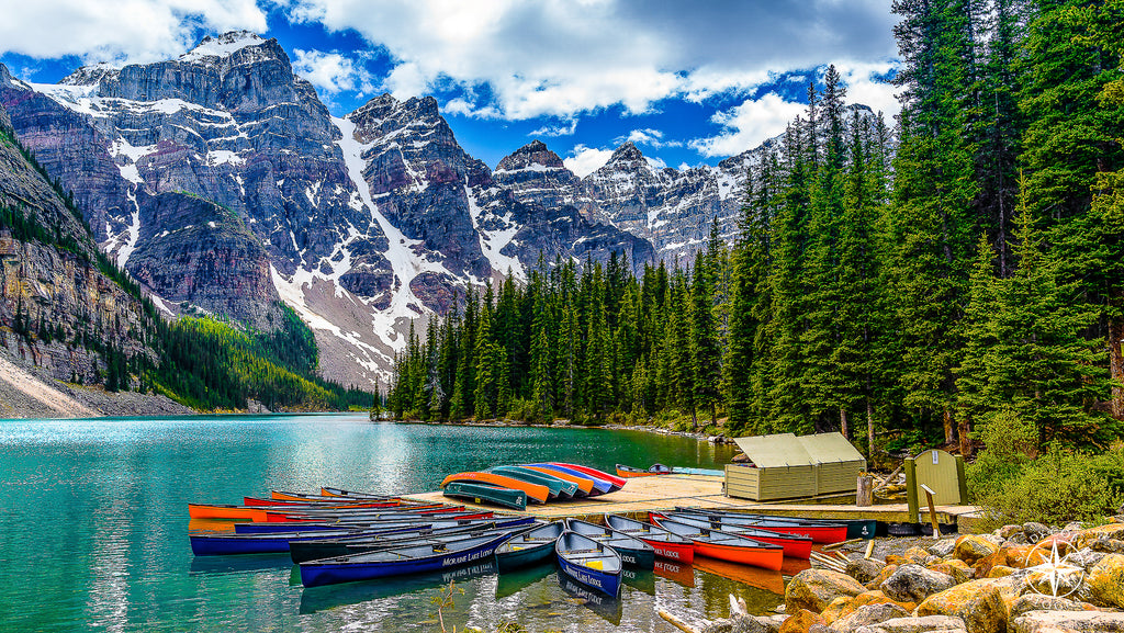A rainbow of canoes fans out from a dock on Moraine Lake in Banff National Park. The teal glacial water sparkles beneath a vi
