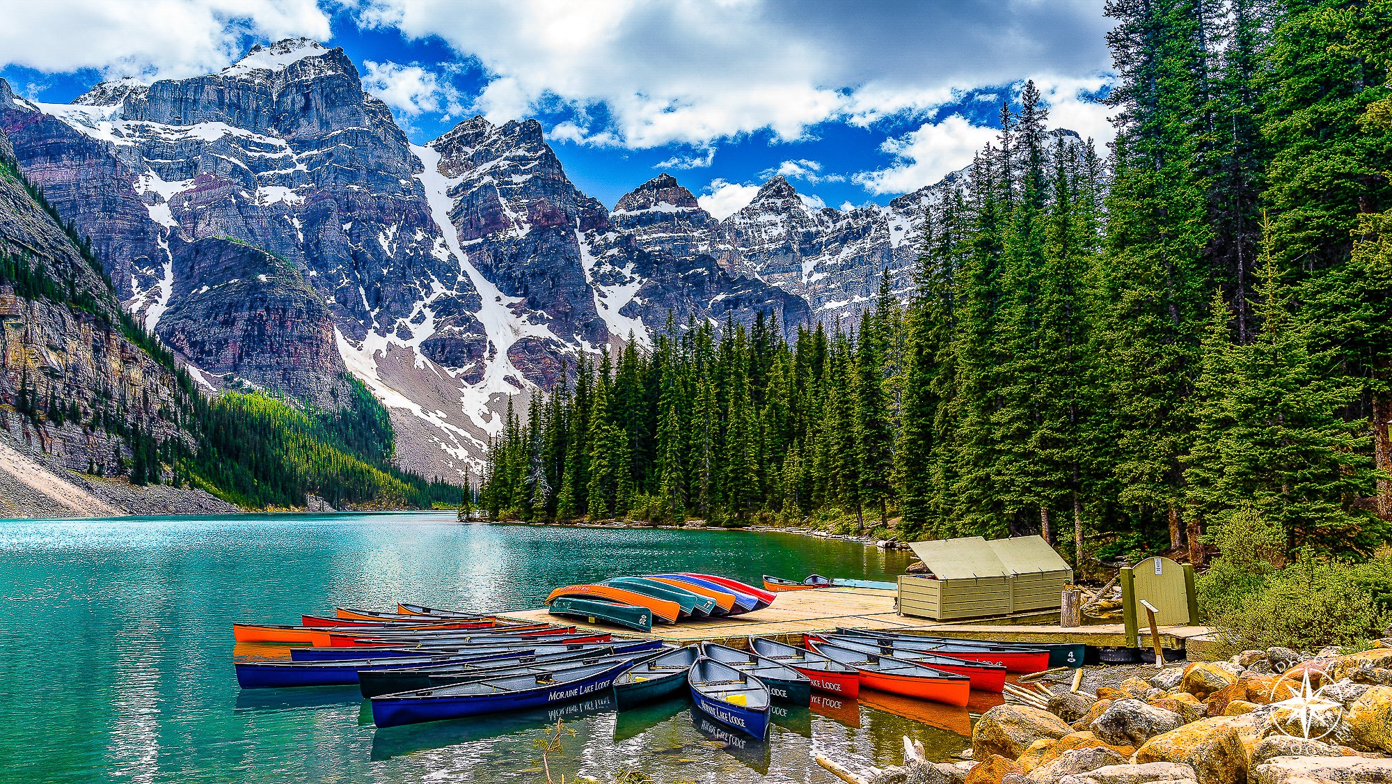 A rainbow of canoes fans out from a dock on Moraine Lake in Banff National Park. The teal glacial water sparkles beneath a vi