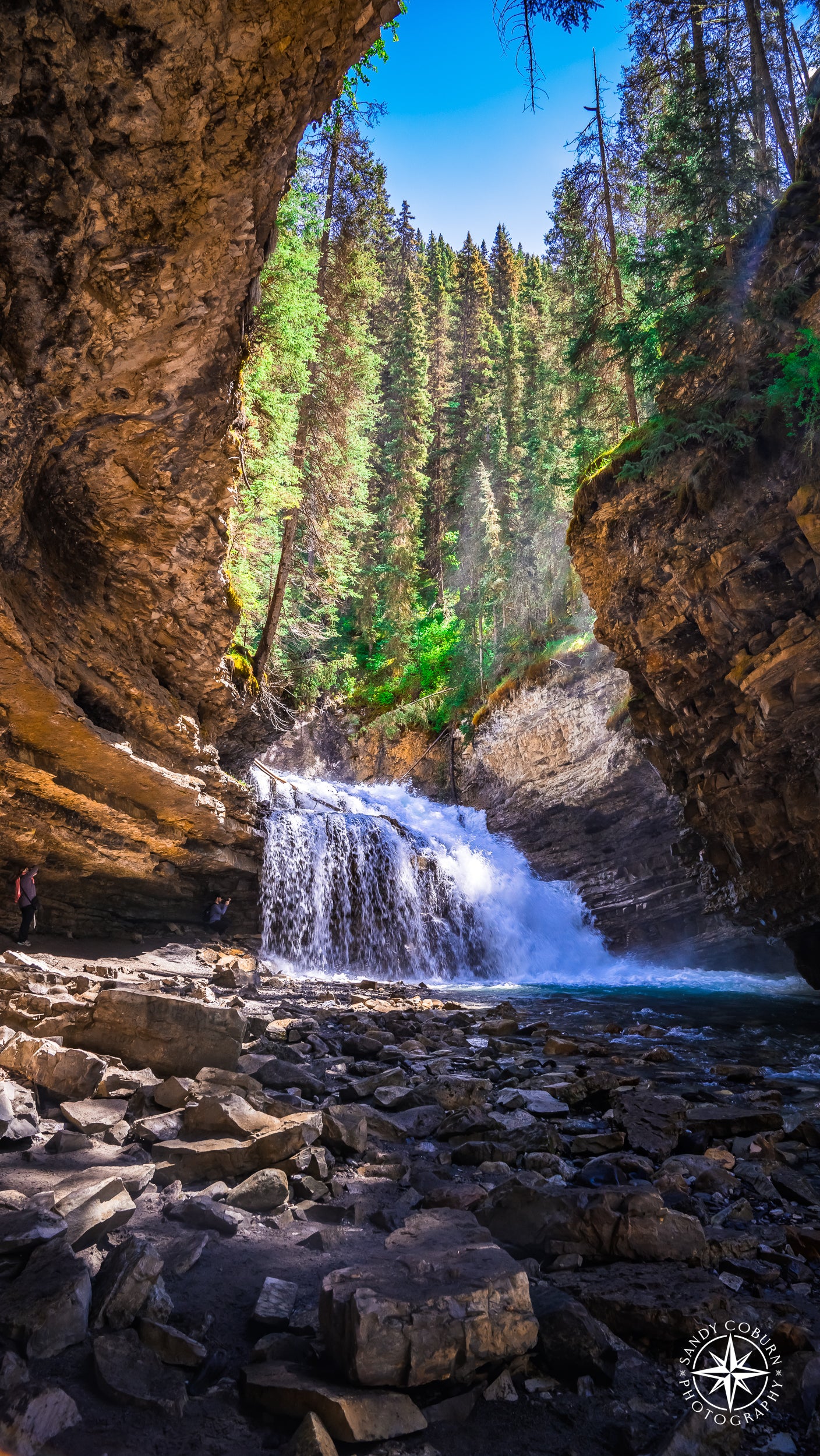 Waterfall framed by curved canyon light in Johnston Canyon, Banff National Park. A distant figure with a camera stands in the