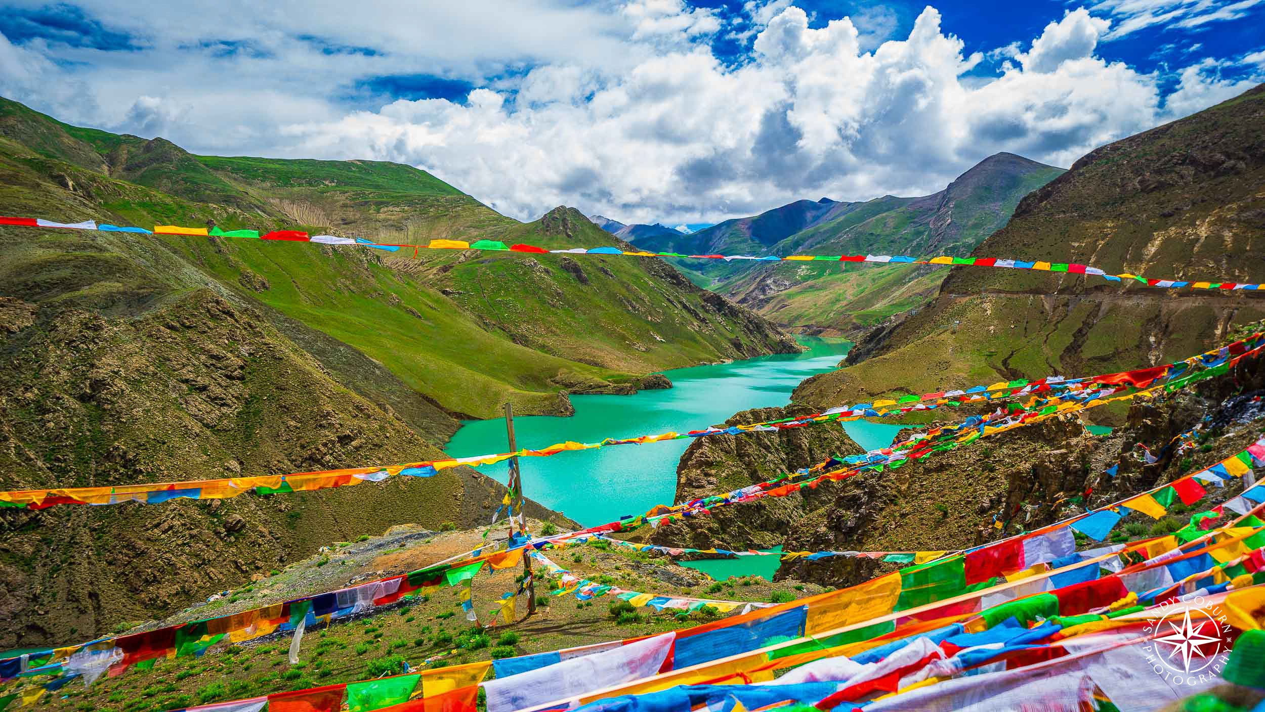 Wide view of the Tibetan Plateau with a dramatic sky, turquoise river, and prayer flags connecting two distant mountains.