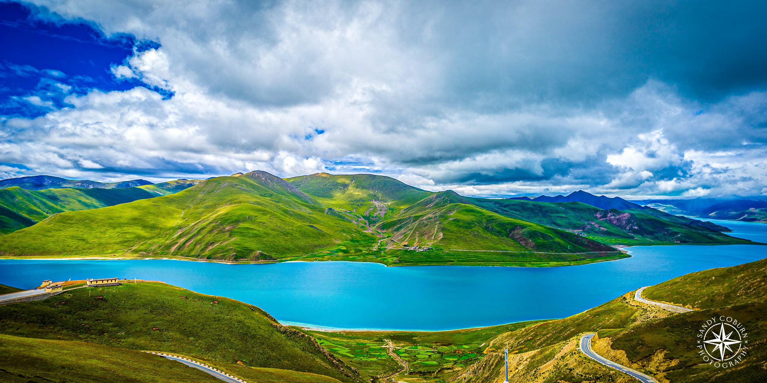 Teal glacial river with a road curving alongside it beneath dramatic clouds on the Tibetan Plateau.