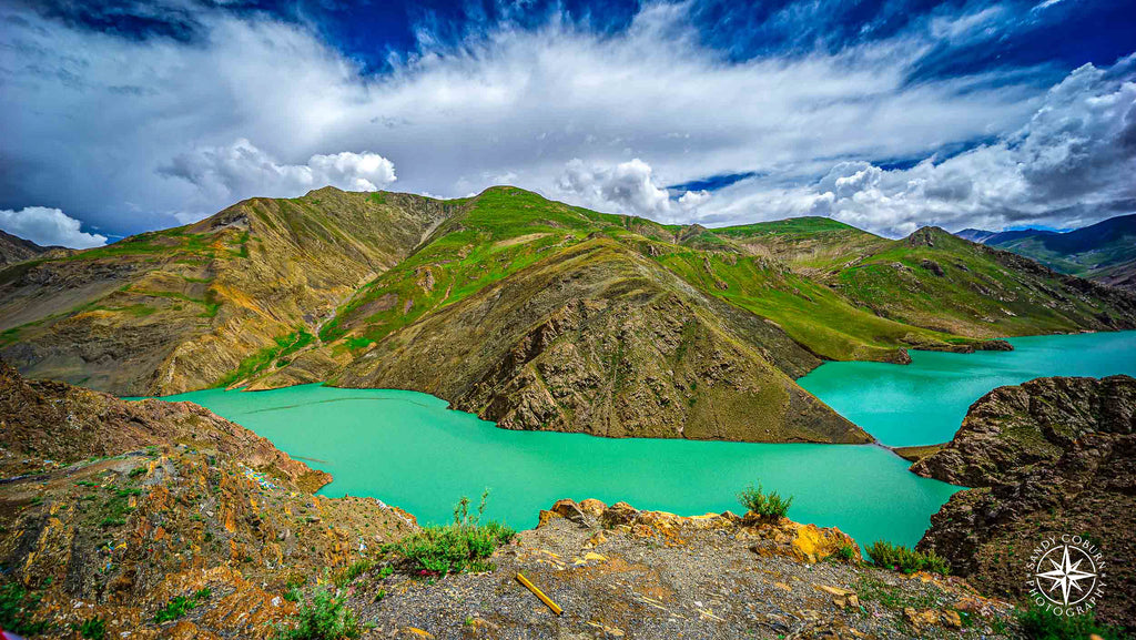 Turquoise river curving through rugged Himalayan terrain with green hills and a soft cloud-filled sky above.