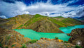 Turquoise river curving through rugged Himalayan terrain with green hills and a soft cloud-filled sky above.