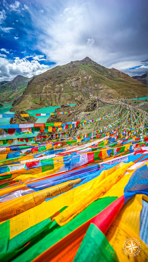 Colorful prayer flags arc across a deep river canyon on the Tibetan Plateau, connecting two distant mountains under a dramati